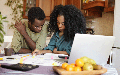 A man and woman sit together looking over paperwork on the table in front of them. There is a laptop computer and a bowl of citrus fruits.