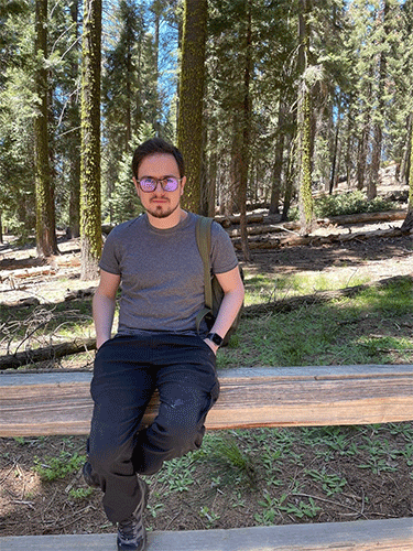 Pedro V. Staziaki sitting on a stone wall with a forest in the background.