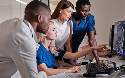 A group of radiologists and radiology technicians are standing and sitting in front of a monitor displaying a radiology image