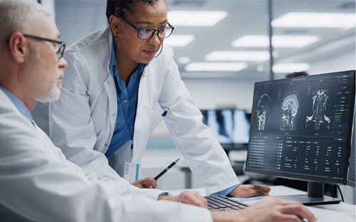 Two medical professionals each wearing glasses and lab coats view a radiology image on a monitor