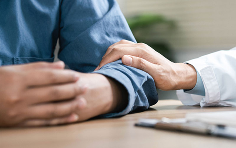 The hand of someone wearing a white lab coat rests on the arm of someone, a patient, wearing a blue shirt