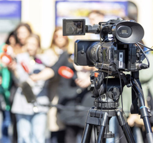 In focus news camera points at a group of people out of focus with microphones. 