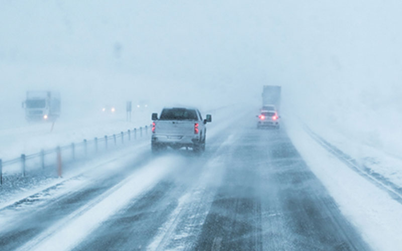 The view of cars and a truck ahead from the driver's seat of a car on a snowy road. 