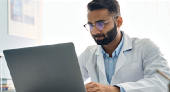 Man wearing glasses and a whitecoat looks at a laptop screen.