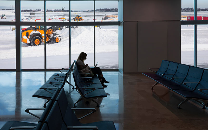 A woman sits in an empty airport terminal, working on her laptop, with windows in the background. Snow is on the ground and airplanes are parked.