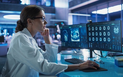 A female radiologist wearing glasses and a white lab coat sits in front of several monitors looking at brain imaging