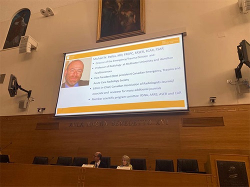 Large screen displays image of Michael N. Patlas MD, listing accomplishments, and hangs above a long desk where panel members are seated