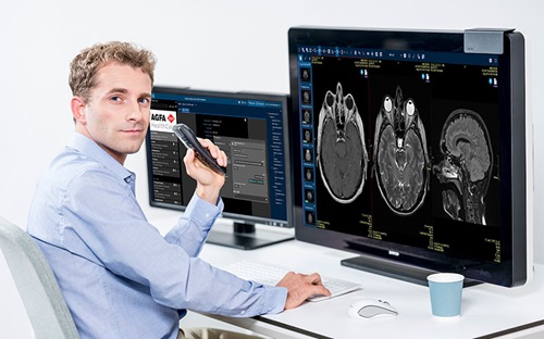 A radiologist is looking over his shoulder at the camera as he sits at a table in front of monitors displaying medical imaging