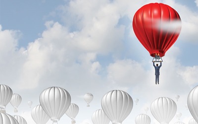 A red hot air balloon piloted by a single person floats in the sky above similar balloons colored only in black and white