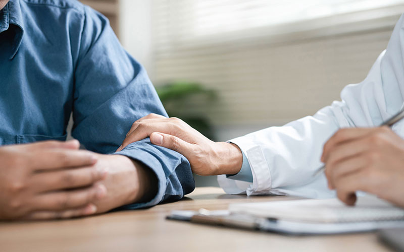 Two people seated at a table with only their arms and the tabletop in view. One, a doctor, in a white coat rests her hand on the arm of the other, a patient, wearing a blue dress shirt