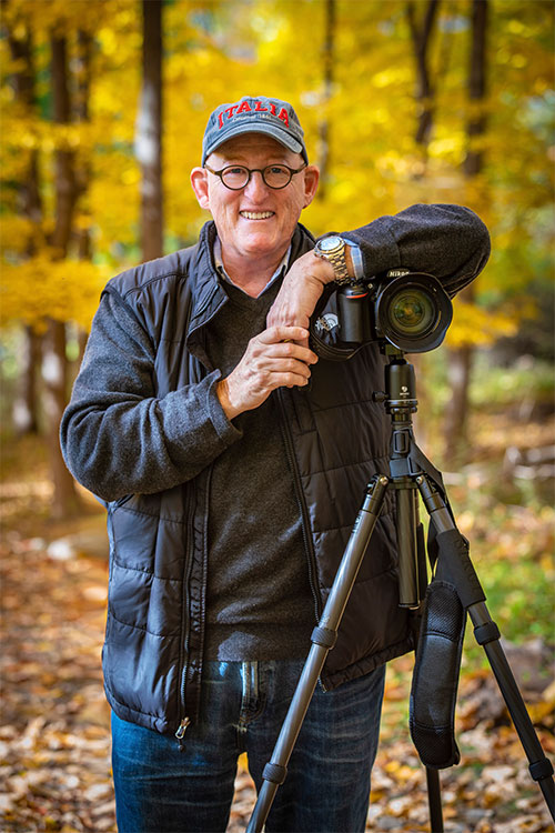 RSNA Member Spotlight Harry Agress, Jr., MD, stands in a wooded area while leaning on his camera that is on a tripod. 