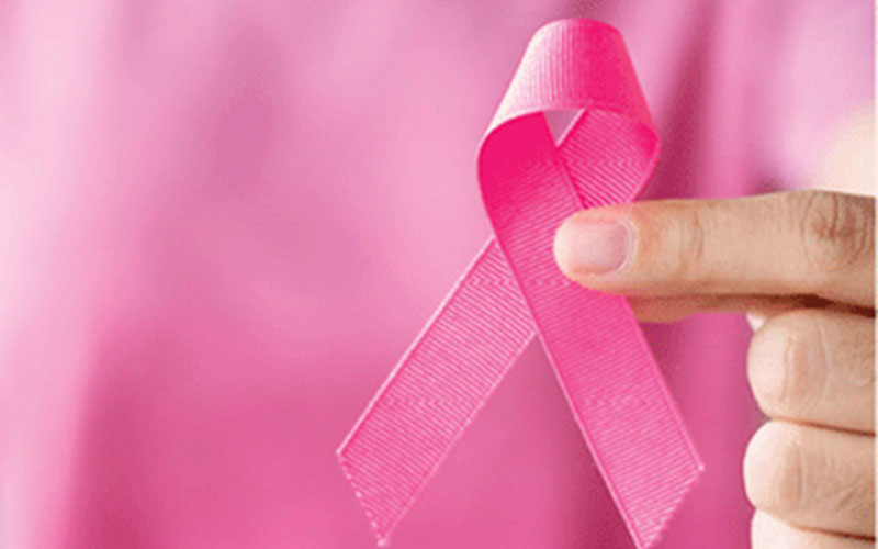 The hand of a woman holding a pink breast cancer awareness ribbon against a pink background