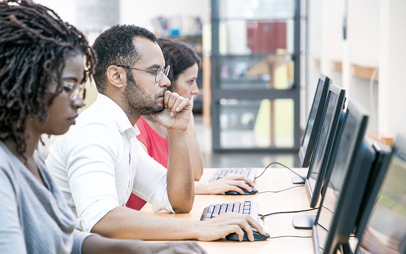 Three students of varying ethnicities sit at a table working on computers.
