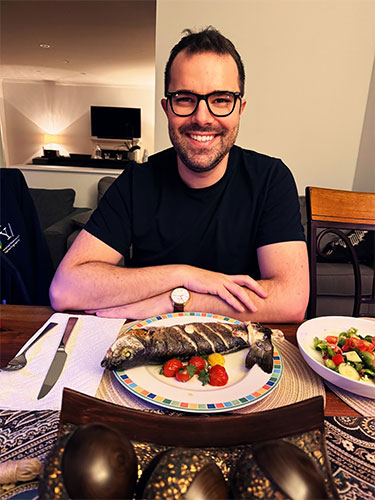 Julius Chapiro, MD, PhD, wearing glasses and a dark t-shirt sits at a table with a plate of fish and vegetables on plate in front of him. 