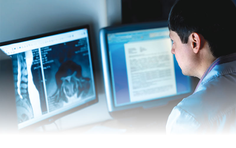 Male radiologists sits looking at two computer monitors, one with images and one with text 