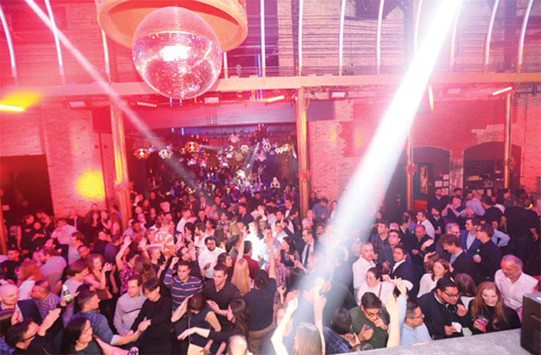 Crowd gathered on a dance floor at an indoor event venue, illuminated by bright stage lights and red ambient lighting. A large disco ball hangs from the ceiling above attendees, with exposed brick walls and industrial-style architecture visible in the background.