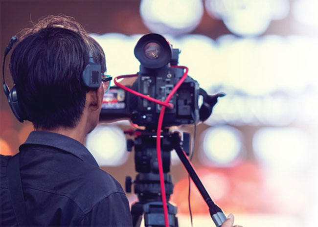 Person operating a professional video camera mounted on a tripod, viewed from behind, with blurred lights in the background.