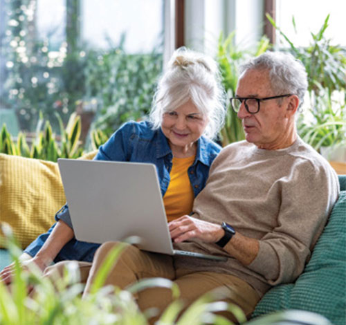 An elderly man and woman sit on a couch and look at a computer. 