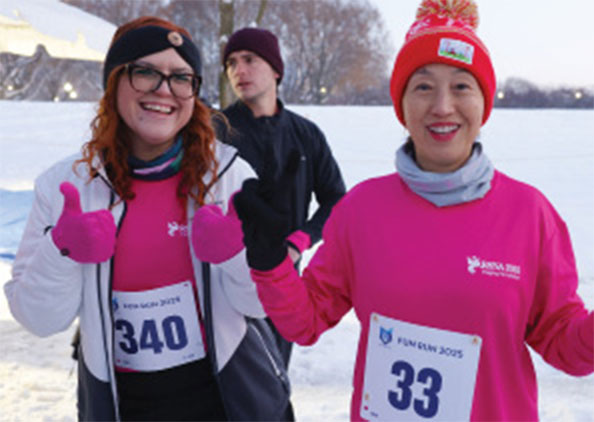 Two participants in bright pink shirts and winter hats gesture toward the camera at a snowy 5K event.