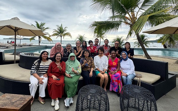 Large group of people on a patio pose for a picture with water behind them and palm trees an patio umbrellas around them.