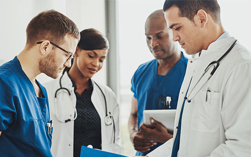 Four people, two wearing scrubs and two wearing lab coats and stethoscopes, stand together looking down at a file folder.