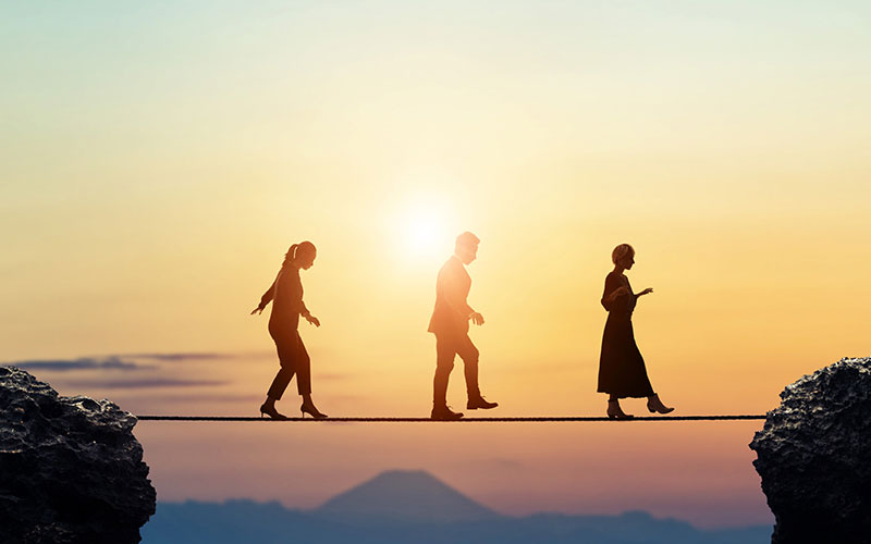 Silhouette of three people who are dressed professionally walking a tightrope between two rock formations with the setting sun in the background.