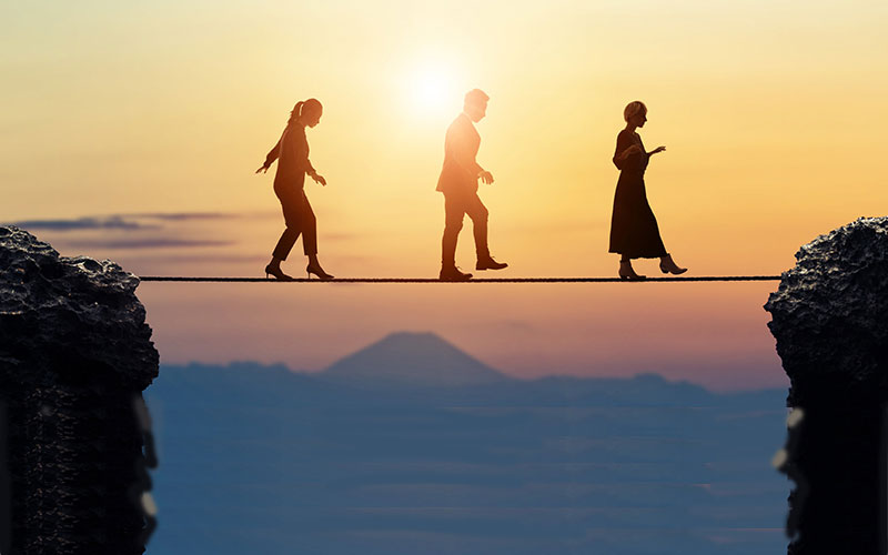 Three people dressed in professional clothing walk a tightrope between 2 rock formation with the sun setting in the background