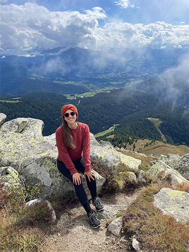Nadine Bayerl, MD, seated on rocks overlooking a mountainous valley with clouds, forests and distant fields below.