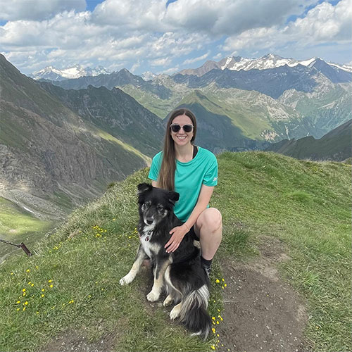 Nadine Bayerl, MD, seated on a grassy mountain ridge with a black-and-white dog, with snow-capped peaks and clouds in the background.