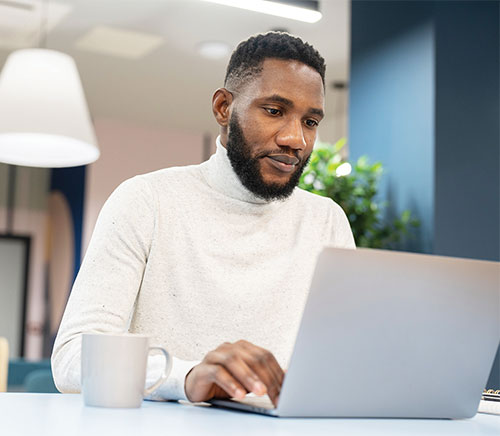 Man with a beard is wearing a white turtleneck shirt and working at a desk on a laptop