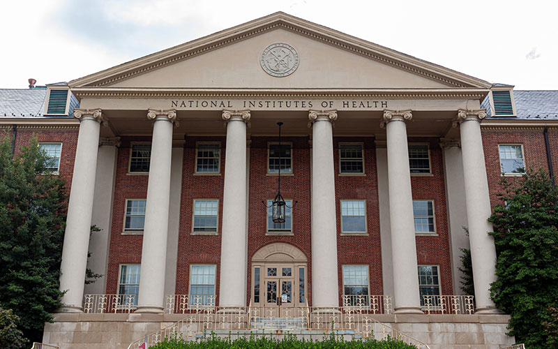 Front entrance of the National Institutes of Health building, featuring a red brick facade with six white columns and the words "National Institutes of Health" above the entrance.