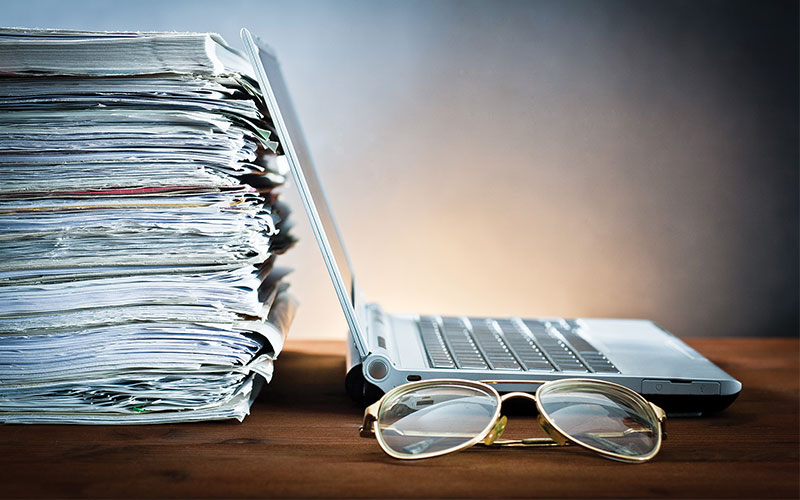 A closeup of a stack of papers at the left with a laptop to the right and a pair of glasses at front, all on a wooden tabletop