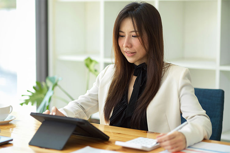Woman sits at a desk holding a pen with a laptop and calculator on the desk in front of her.