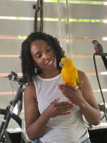 Melissa Davis, MD, MBA, in a sleeveless black-and-white striped top holding a bright yellow parrot indoors; a bicycle, window blinds and a second bird perched in the background.