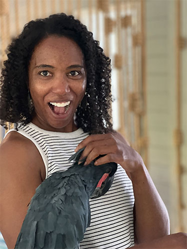 Melissa Davis, MD, MBA, in a sleeveless black-and-white striped top holding a dark gray bird with a red marking near its head indoors, with light wood slats in the background.