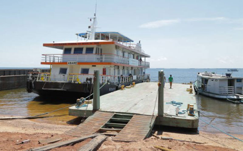 A large boat used for mobile imaging is docked. A man is standing on the dock, and a smaller boat is tied to the opposite side of the dock.
