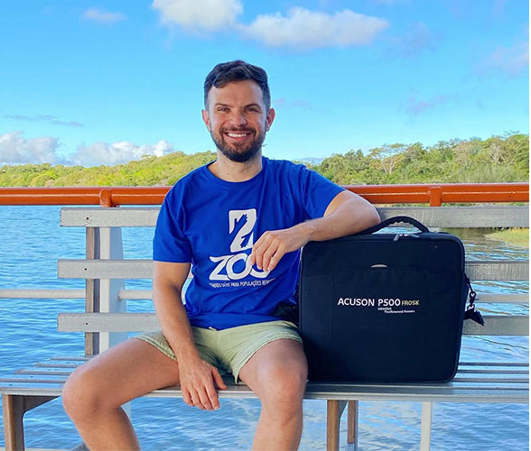 Jose Araujo-Filho, MD, PhD, sits on a bench near water with his arm propped on a duffel bag.