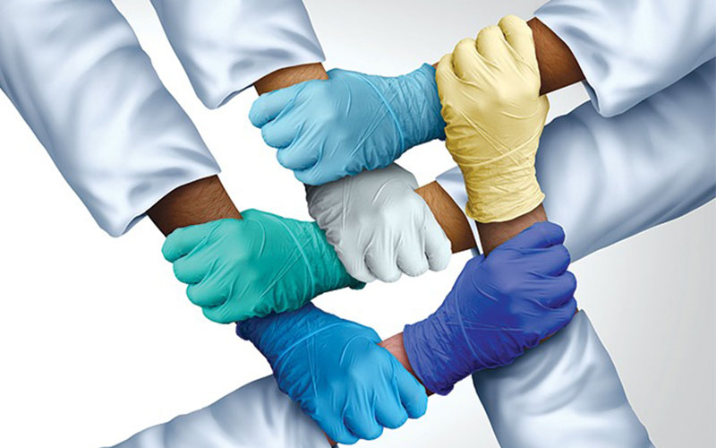 The hands of six people in lab coats wearing latex gloves grab the wrists of each other forming a basketweave pattern.