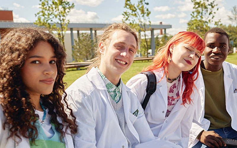 Four young adults sitting outdoors together, three wearing white lab coats, smiling toward the camera on a sunny day with trees and a pergola in the background.