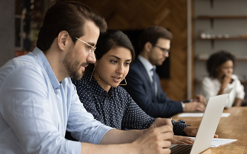Four professionals seated side-by-side at a conference table, focused on laptops and tablets during a collaborative meeting in an office setting.