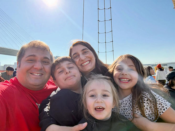 Five people, two adults and three children, taking a close-up outdoor selfie near a suspension bridge on a sunny day.
