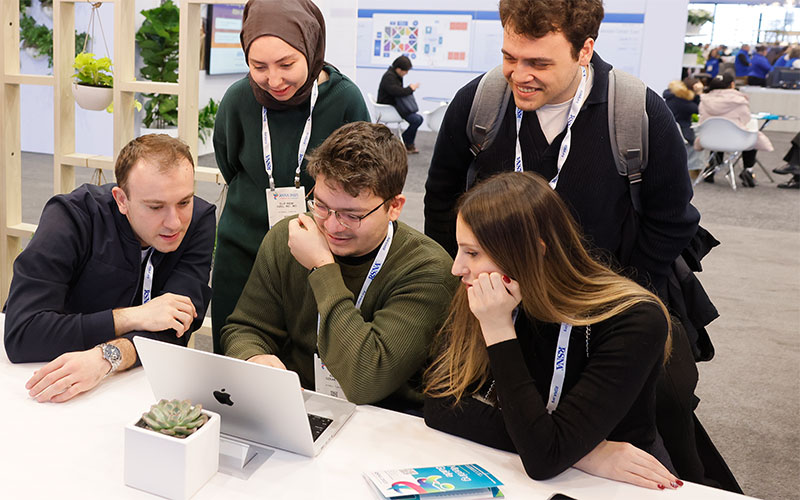 Five attendees wearing conference badges look at a laptop on a table in an open meeting and networking space.