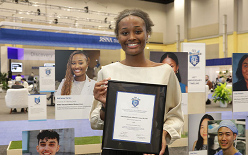 A young woman stands holding a framed certificate in front of a photographic display featuring headshots of other diverse people